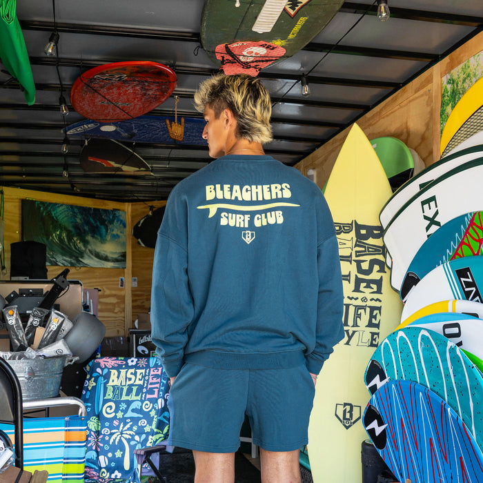 Adult model wearing blue crewneck with "Bleachers Surf Club" design