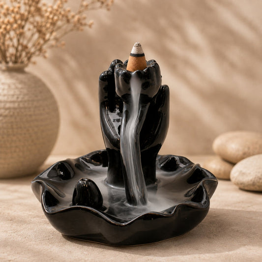 A glossy black ceramic backlit incense burner shaped like two hands holding a smoking incense cone, with smoke cascading down the center into a shallow dish. The product is styled on a beige textured surface with a blurred beige background, a decorative vase with dried stems on the left, and smooth stones on the right.