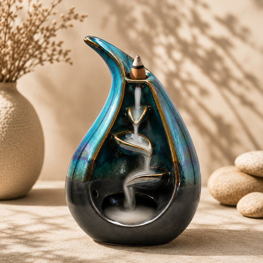 A ceramic teardrop-shaped incense burner with a flowing white incense trail, featuring a blue, green, and black iridescent glaze with gold accents, styled on a beige surface with decorative stones and a vase in the background.