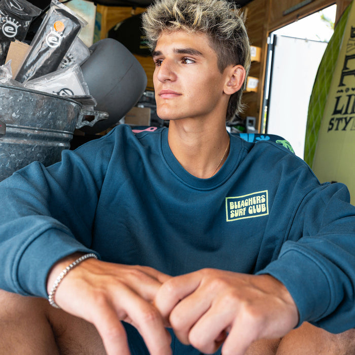 Adult model wearing blue crewneck with "Bleachers Surf Club" design