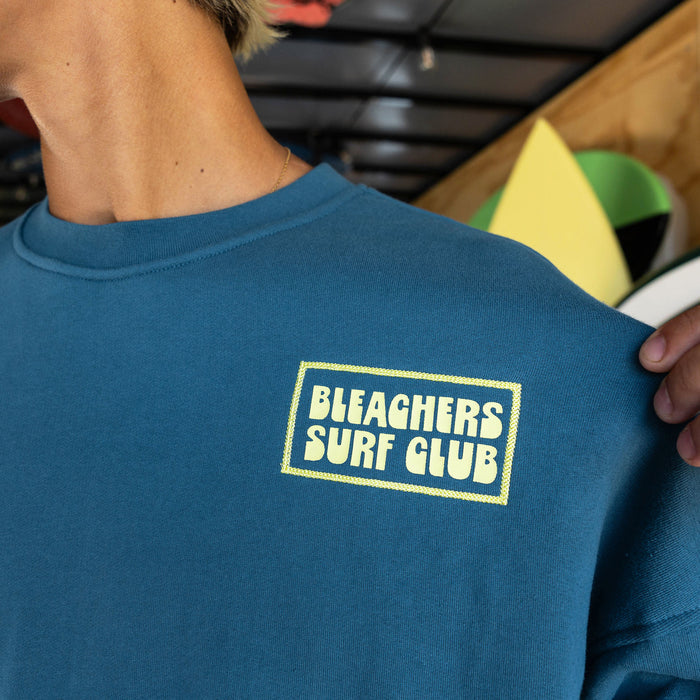 Adult model wearing blue crewneck with "Bleachers Surf Club" design