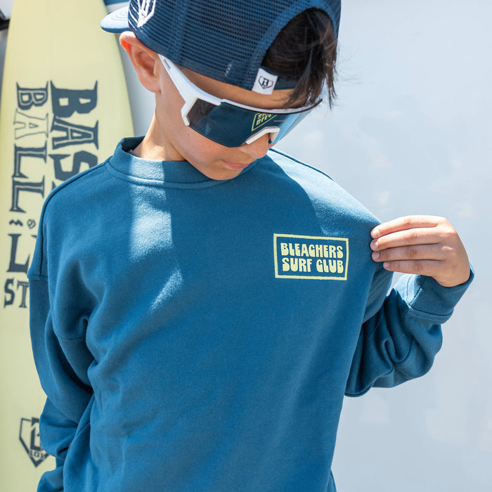 Youth model wearing blue youth crewneck with "Bleachers Surf Club" design