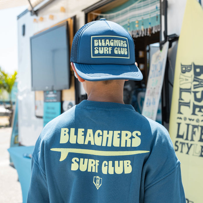 Youth model wearing blue youth crewneck with "Bleachers Surf Club" design