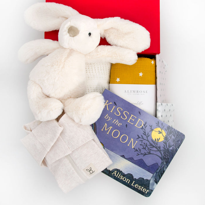 A baby hamper set including a vanilla-colored plush bunny, an oatmeal-colored organic cotton romper, a butterscotch star organic muslin swaddle, and a white cellular blanket, with a children's book titled 'Kissed by the Moon'. Price: 159