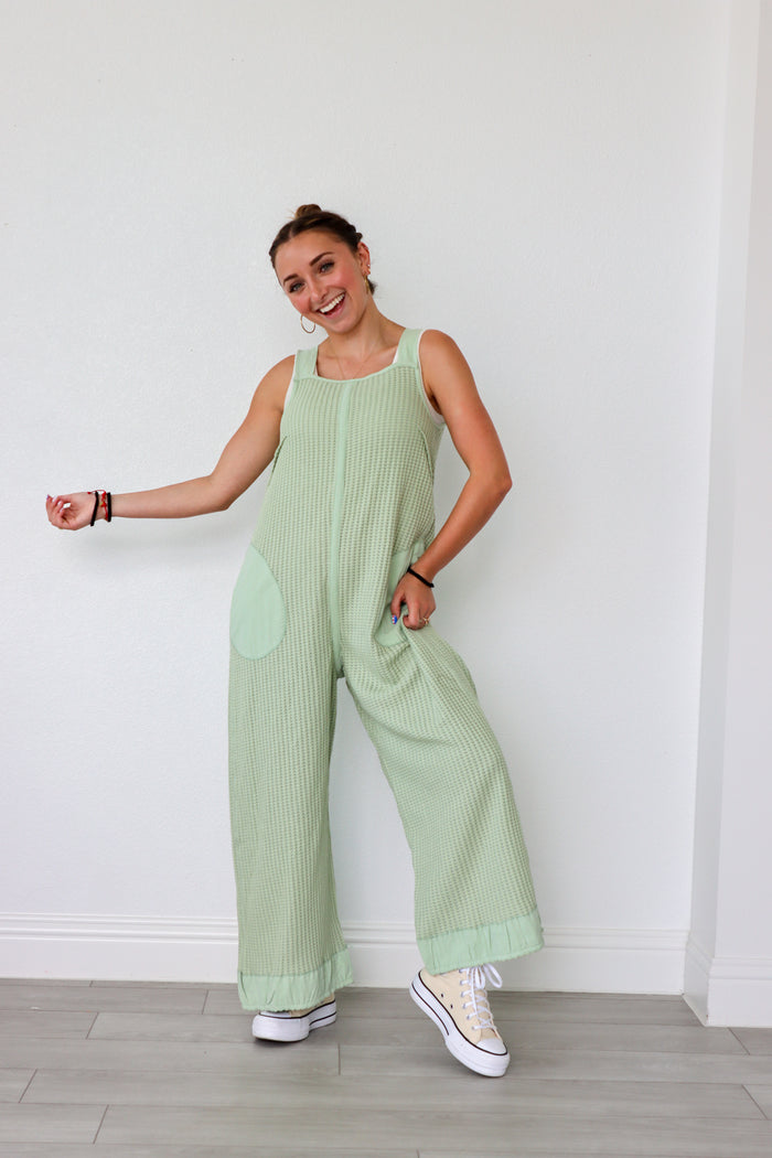 Woman wearing a light green ribbed jumpsuit against a white wall.