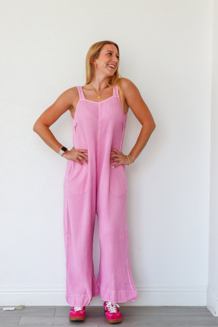 Woman wearing a pink jumpsuit standing against a white wall.
