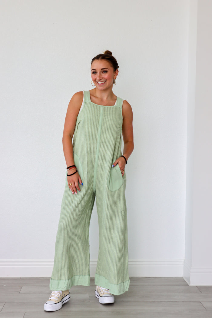 Woman wearing a light green ribbed jumpsuit against a white wall.