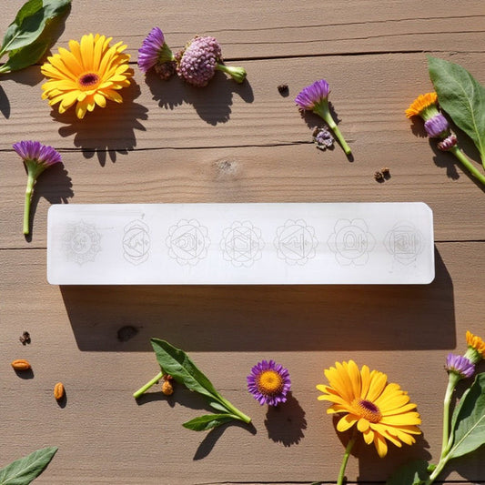 White rectangular selenite bar with chakra patterns on a wooden surface with flowers and leaves.