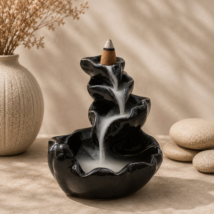 A black ceramic tiered waterfall incense burner with white smoke flowing down the steps, topped with a congee incense stick, set on a beige fabric surface with dried flowers and stones in the background.