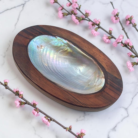 Wooden shell dish with a large shell on a marble surface with pink flowers.